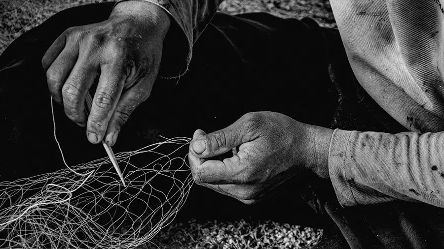 Weathered hands mending a fishing net with a needle. Photograph by Alexandra Martins